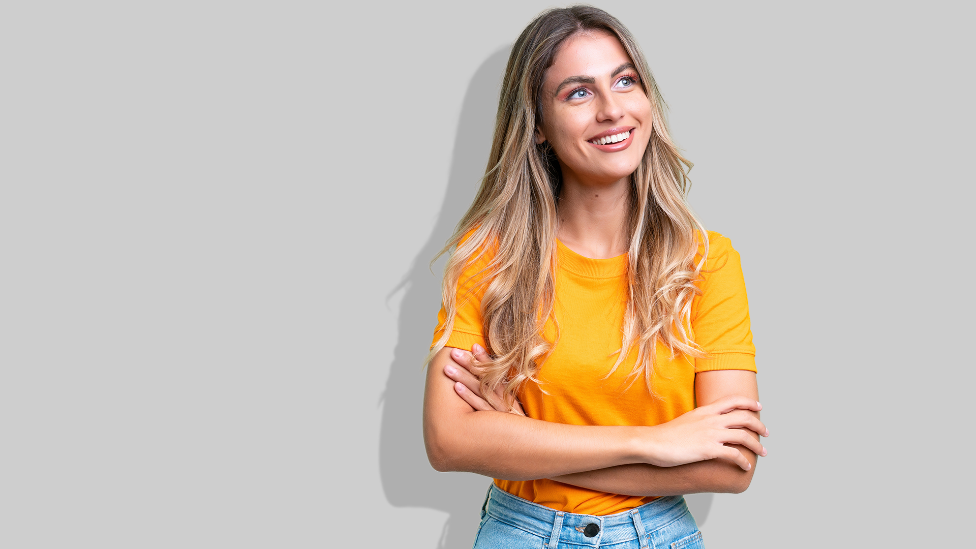 Eine junge Frau mit orangenem T-Shirt und verschränkten Armen steht vor einer grauen Wand und lächelt.