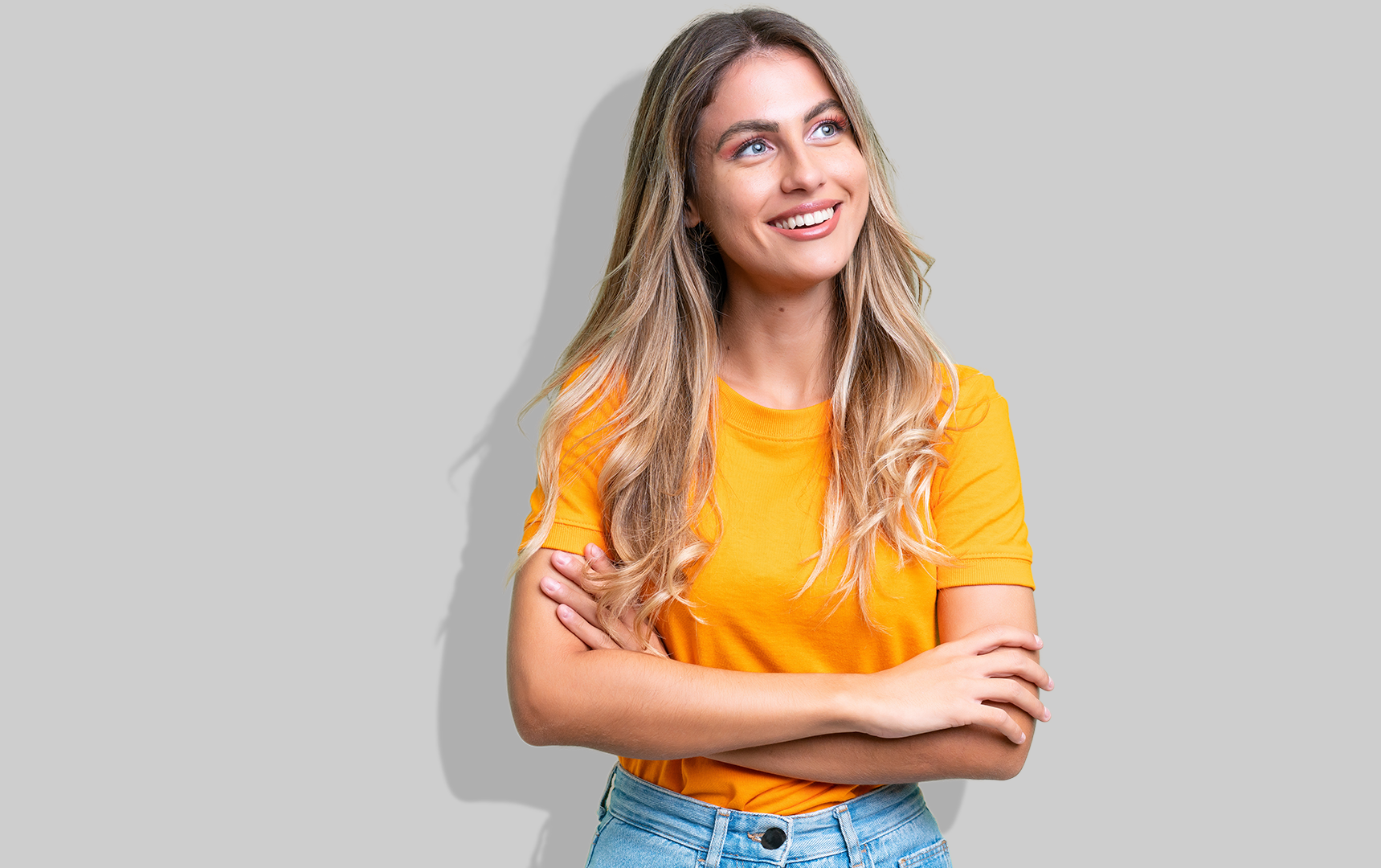 Eine junge Frau mit orangenem T-Shirt und verschränkten Armen steht vor einer grauen Wand und lächelt.