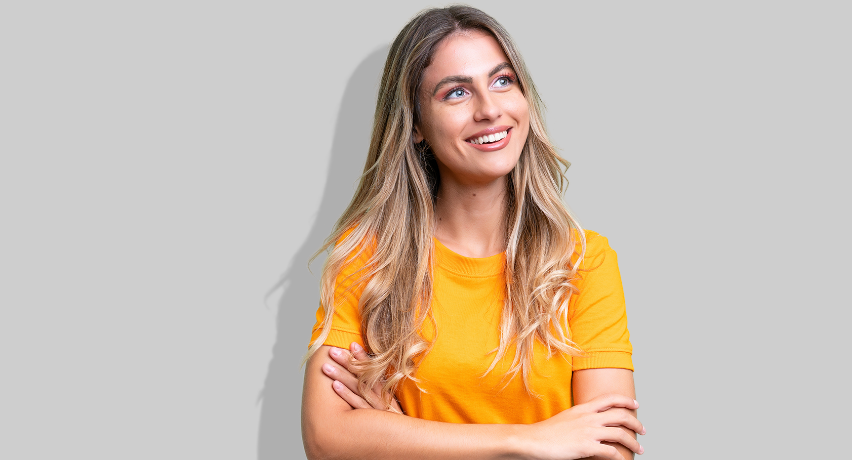 Eine junge Frau mit orangenem T-Shirt und verschränkten Armen steht vor einer grauen Wand und lächelt.