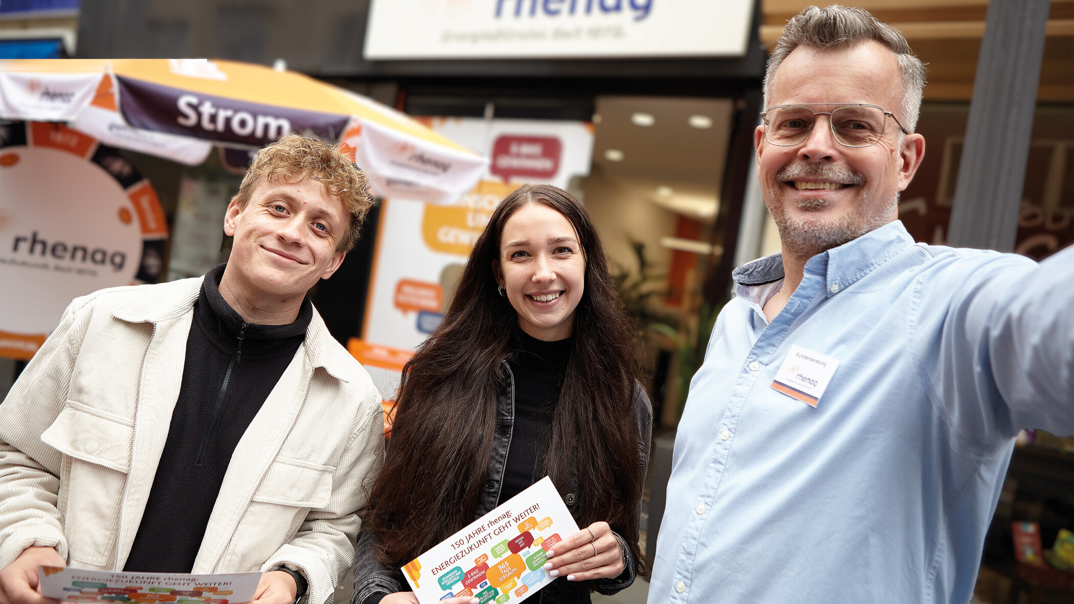 rhenag Mitarbeiter stehen vor dem rhenag Shop und lächeln.