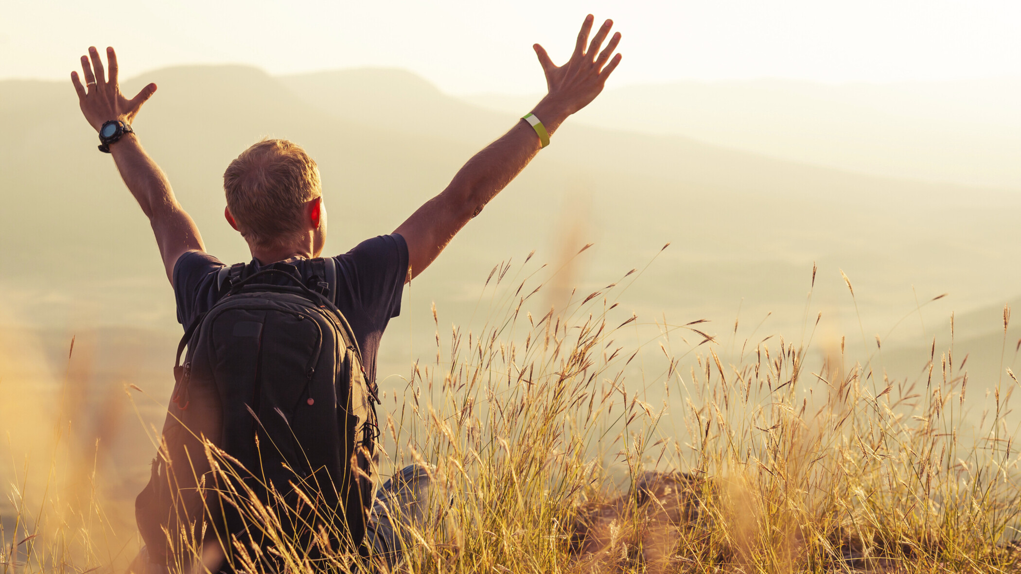 Mann mit Rucksack steht im Feld und freut sich mit erhobenen Armen.